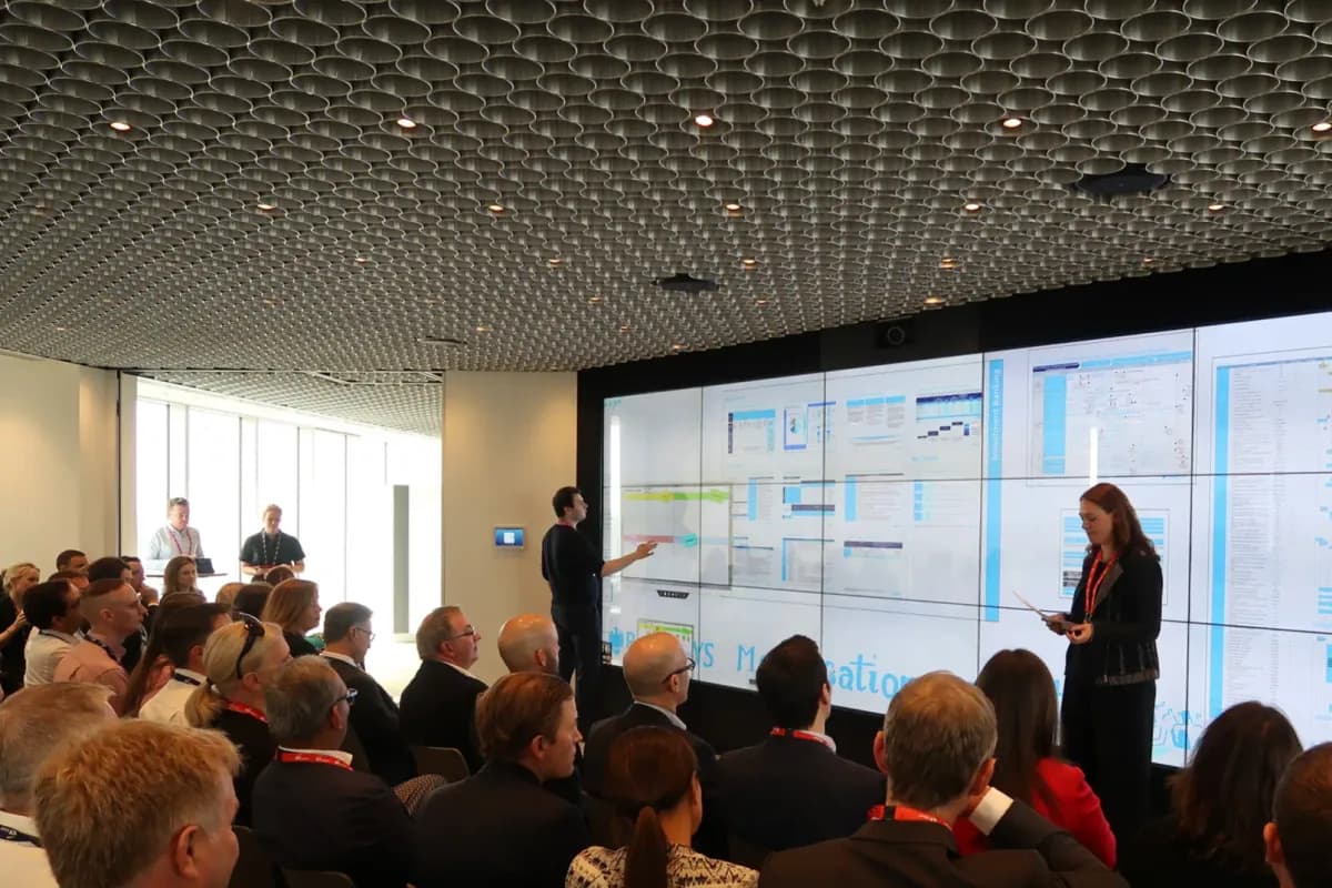 A large-format digital wall at EY showing a grid of documents, with two presenters standing in front of a seated audience.