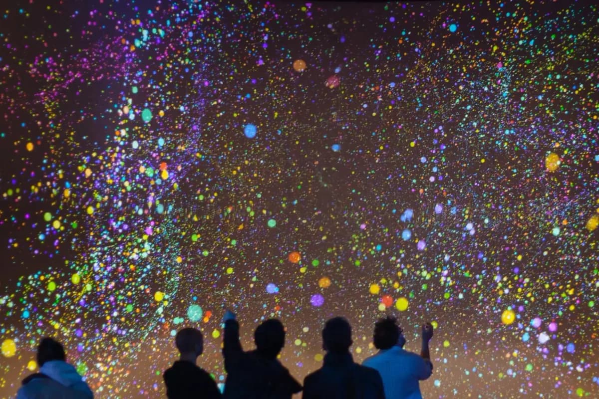 An immersive data visualisation installation: visitors in silhouette reaching towards a wall covered in thousands of coloured dots.