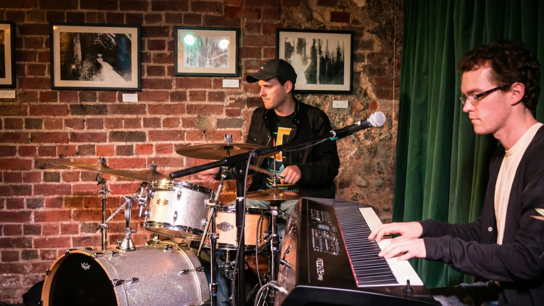 Simon at a silver sparkle drum kit against a bare brick wall in a small venue, with a keyboard player in the foreground.